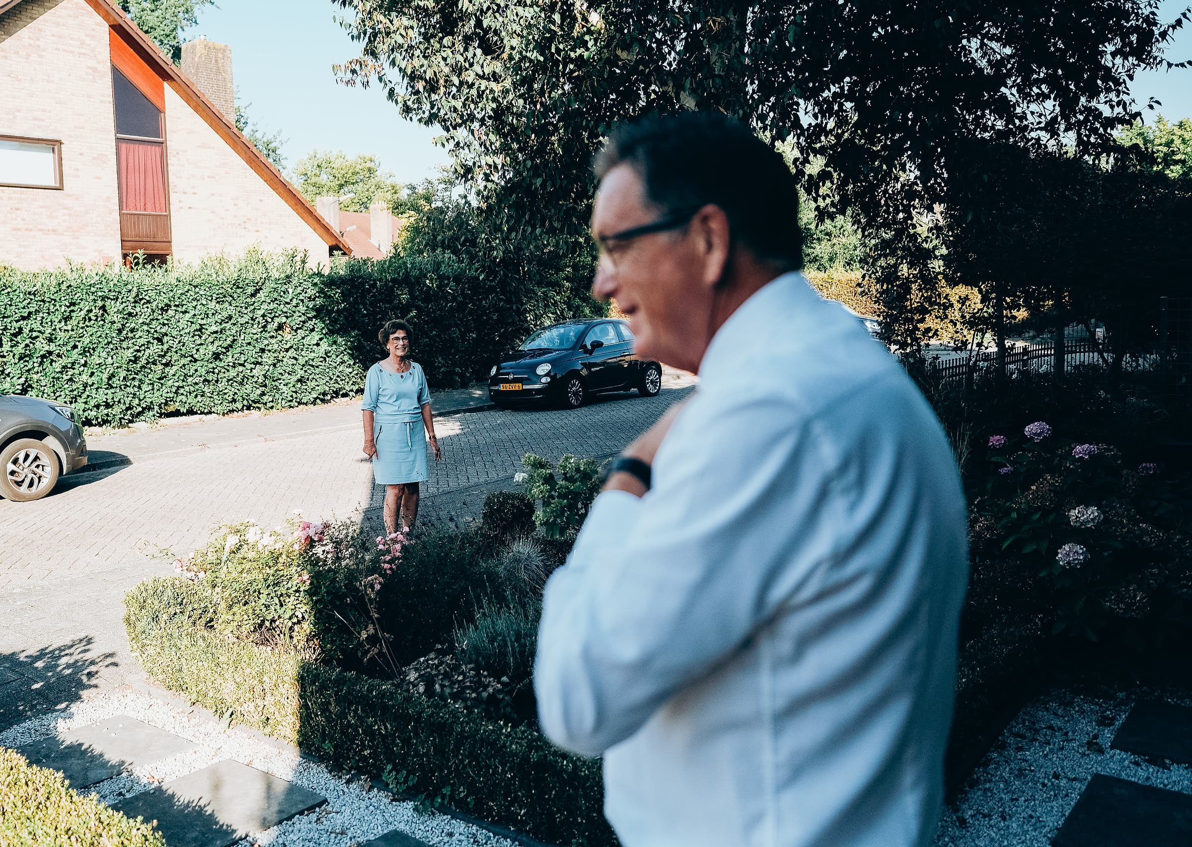 A groom adjusting his cufflink in the foreground while a wedding guest smiles by a hedge in the background