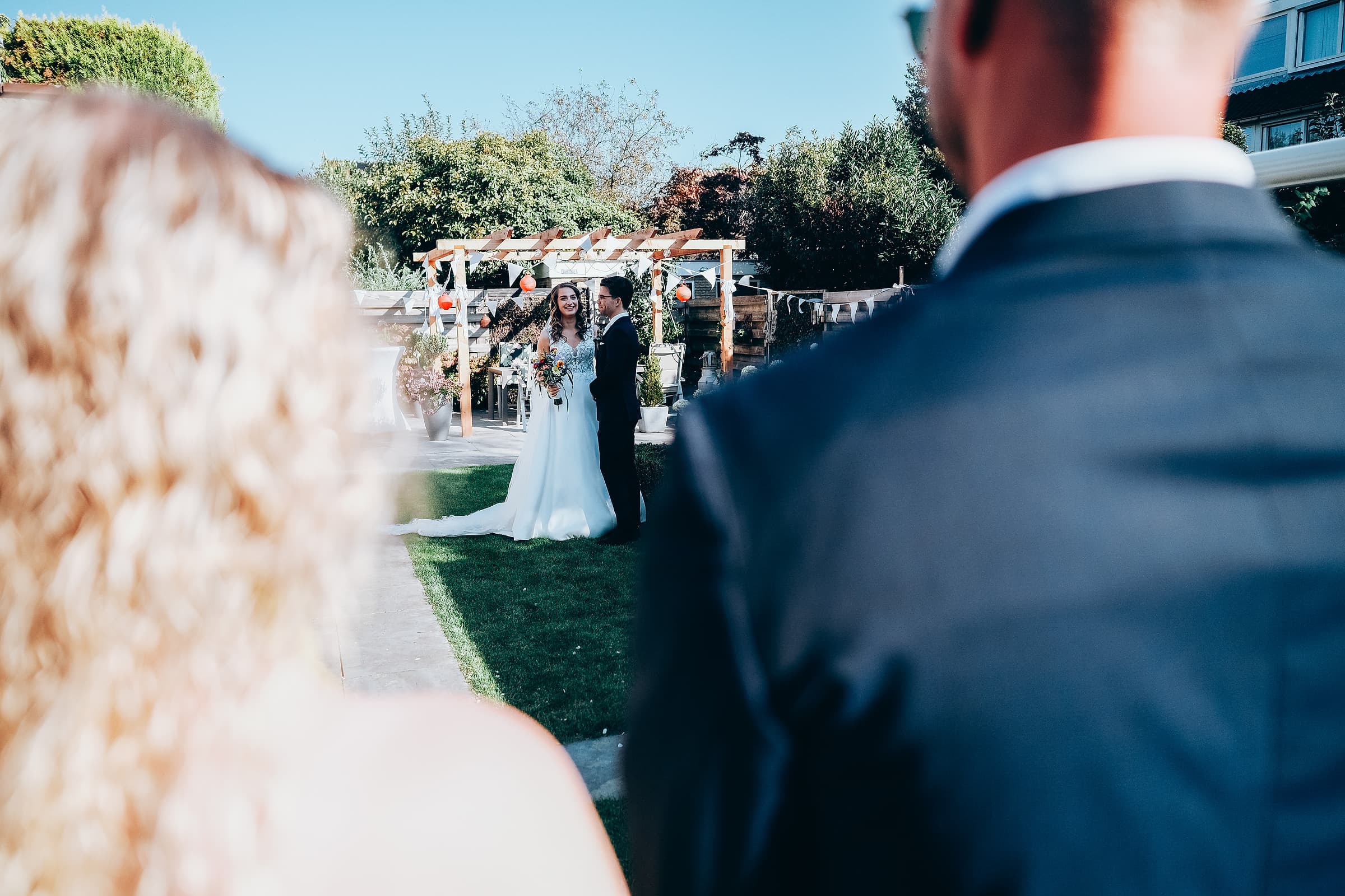 A wedding guest's view over their shoulder as a bride and groom exchange vows in a garden