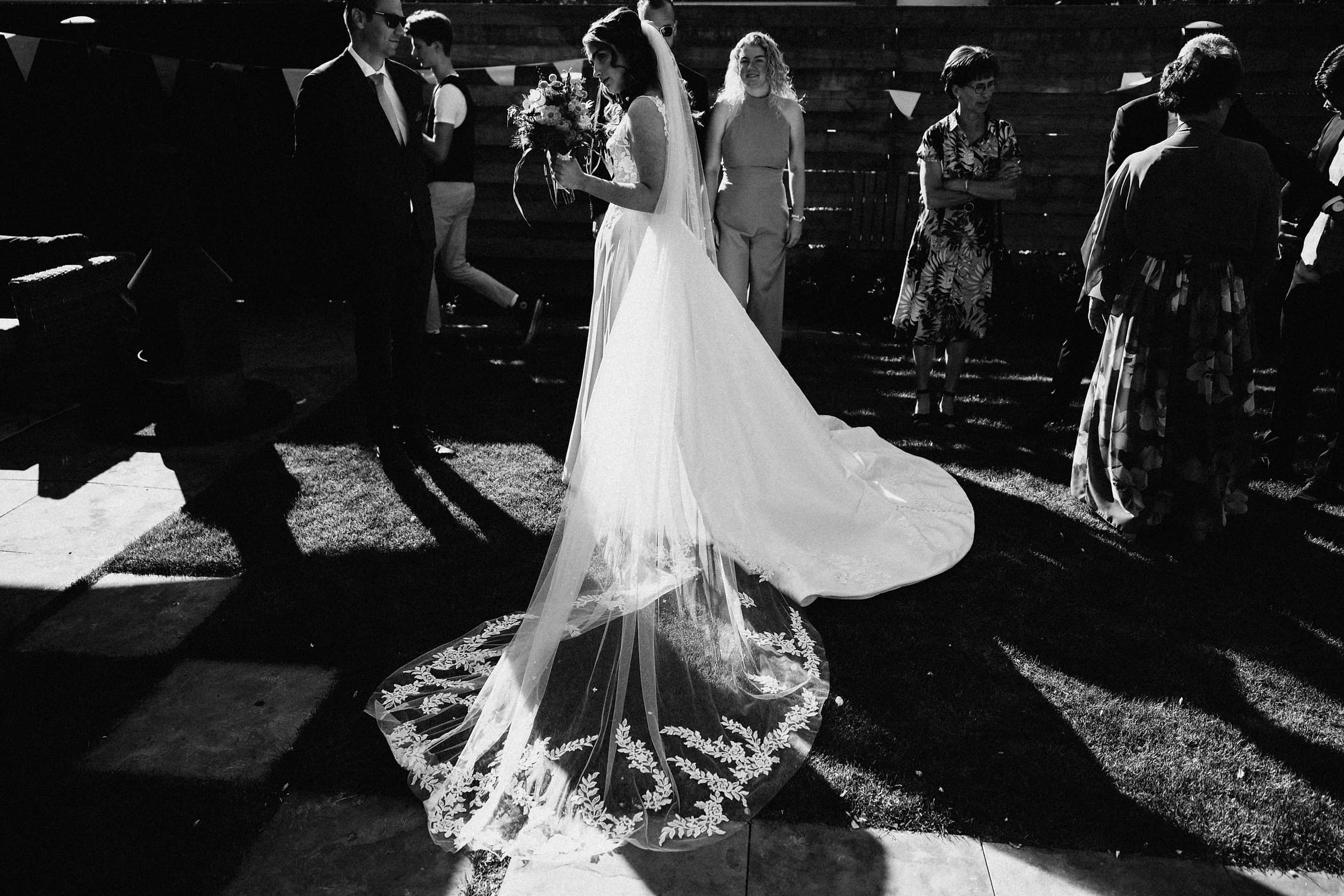 A bride's veil train catching the light as wedding guests look on, in black and white
