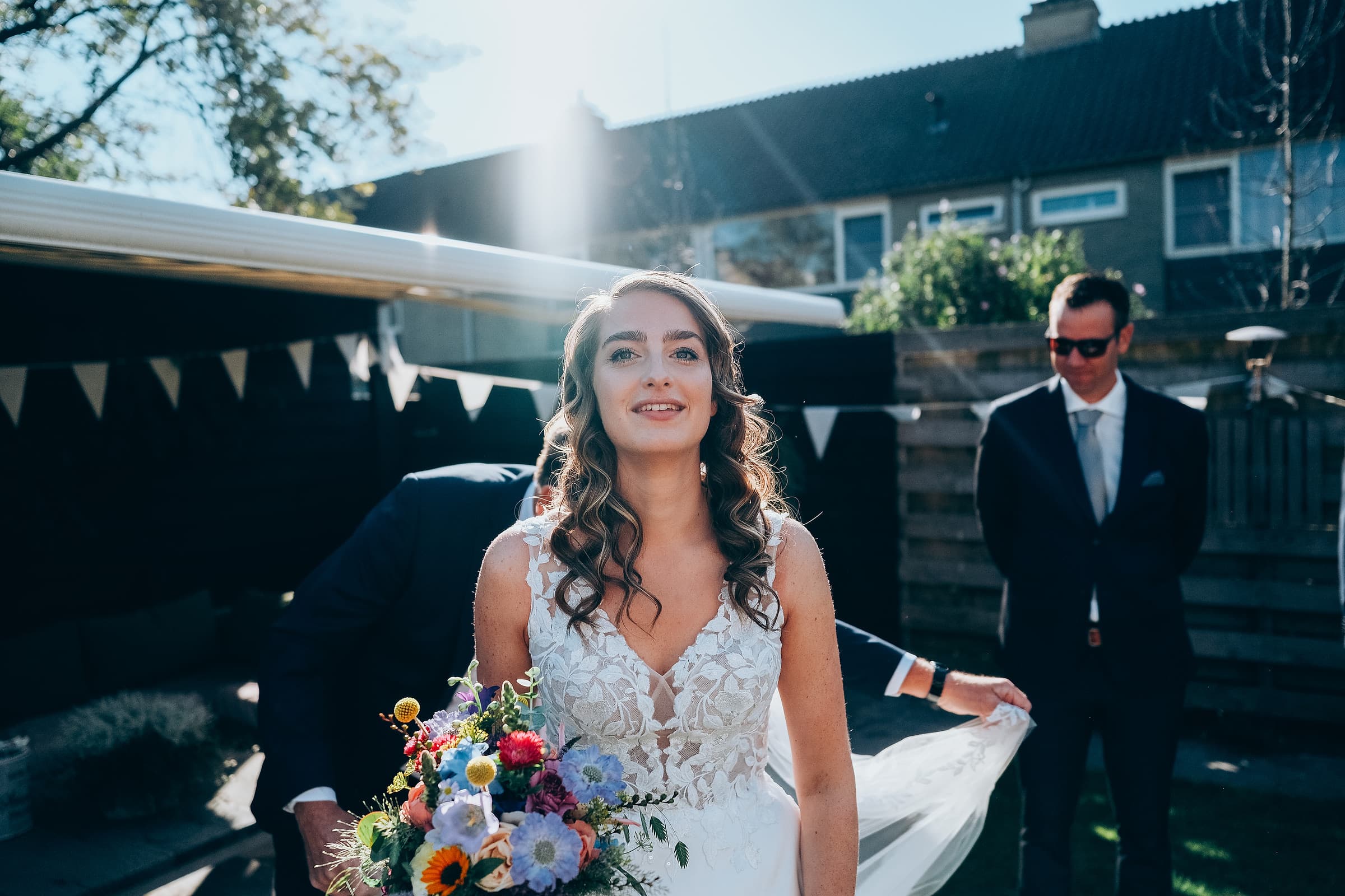 A bride smiling in bright backyard sunlight while holding a colorful bouquet