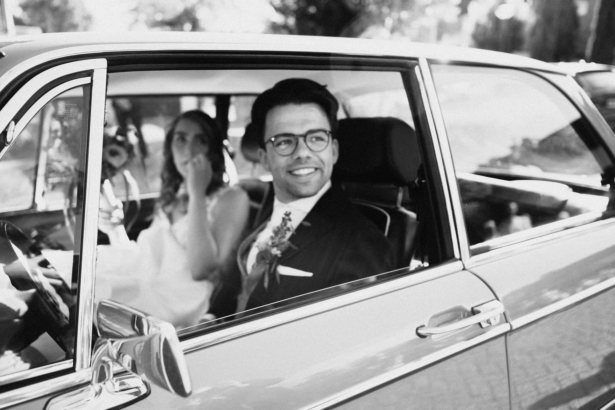A candid black and white photo of a bride and groom laughing together in the back of a vintage car