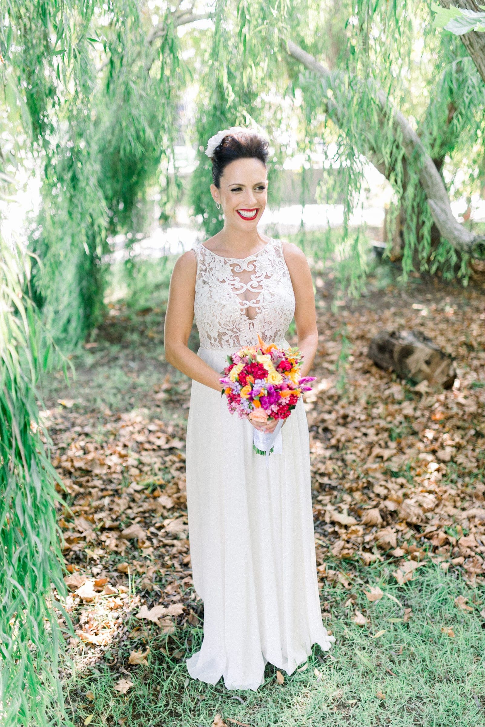 A bride in a lace wedding dress smiling under hanging willow branches, holding a bright bouquet