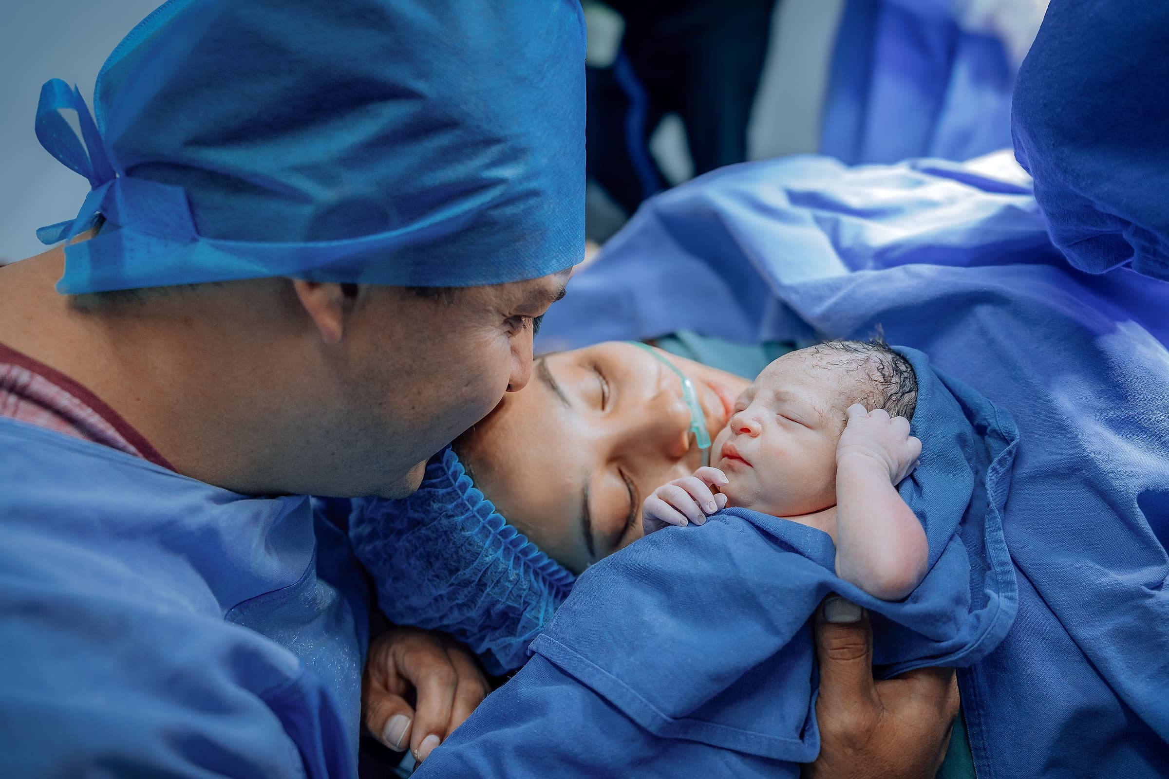 Two parents leaning in to kiss their newborn baby moments after birth in a hospital delivery room