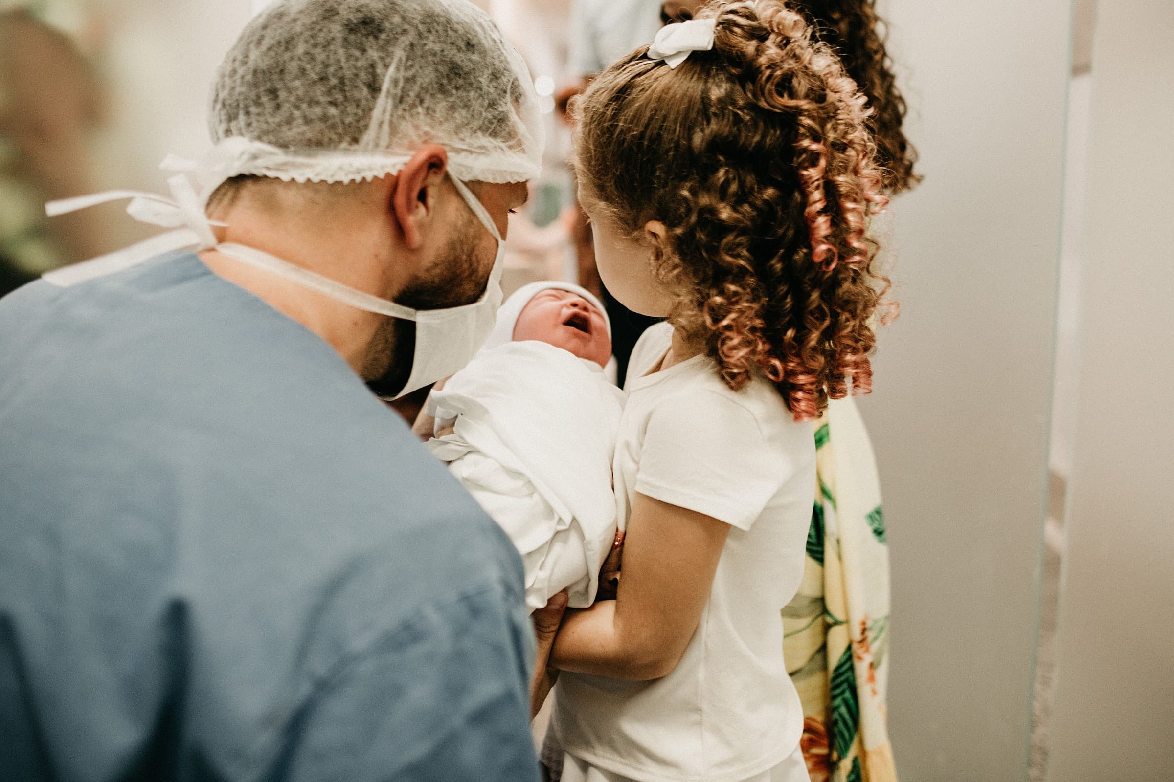 A father in hospital scrubs holding a newborn as an older sister meets her sibling for the first time
