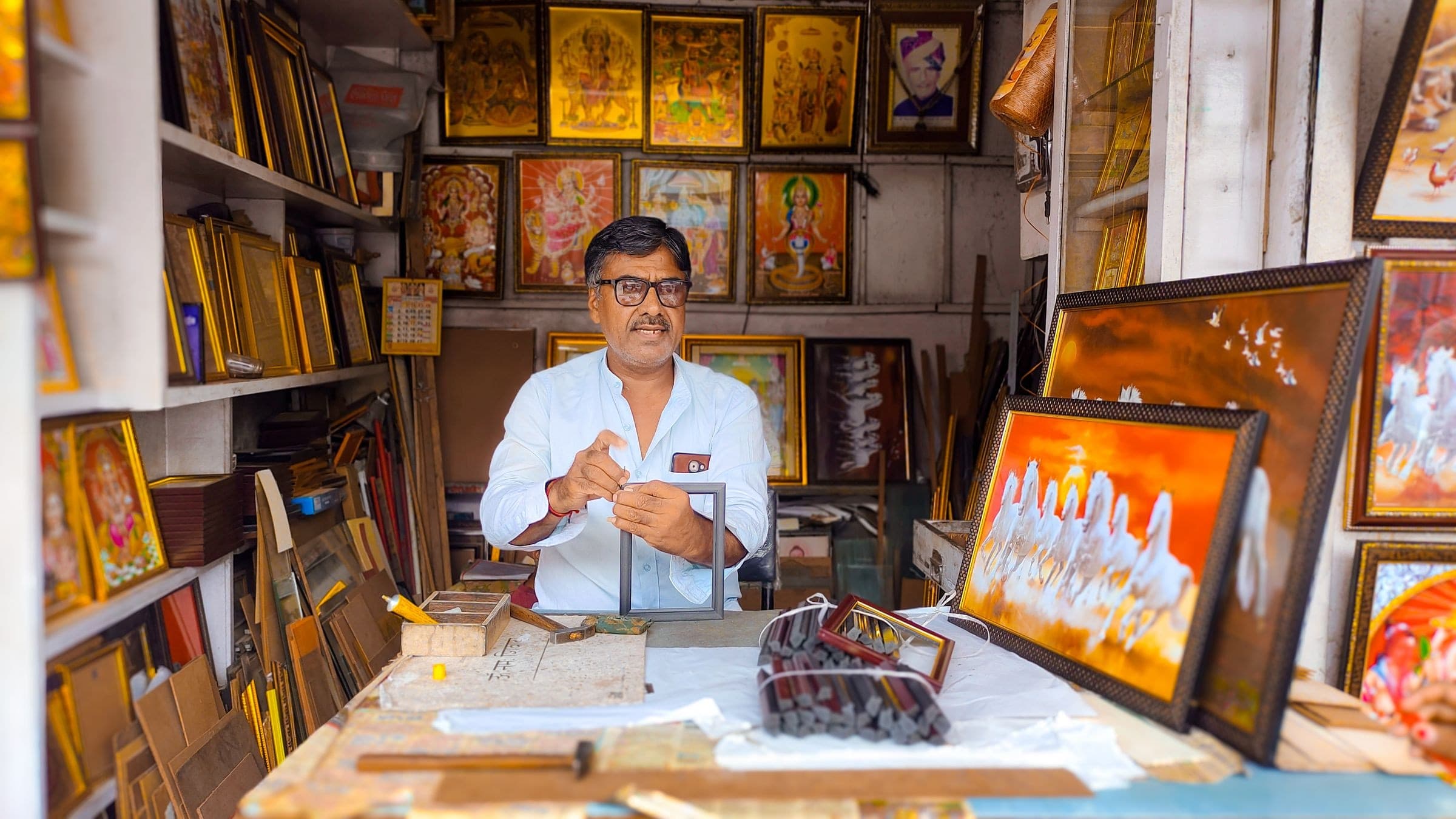 A shop owner assembling a picture frame at his workbench, surrounded by his stock