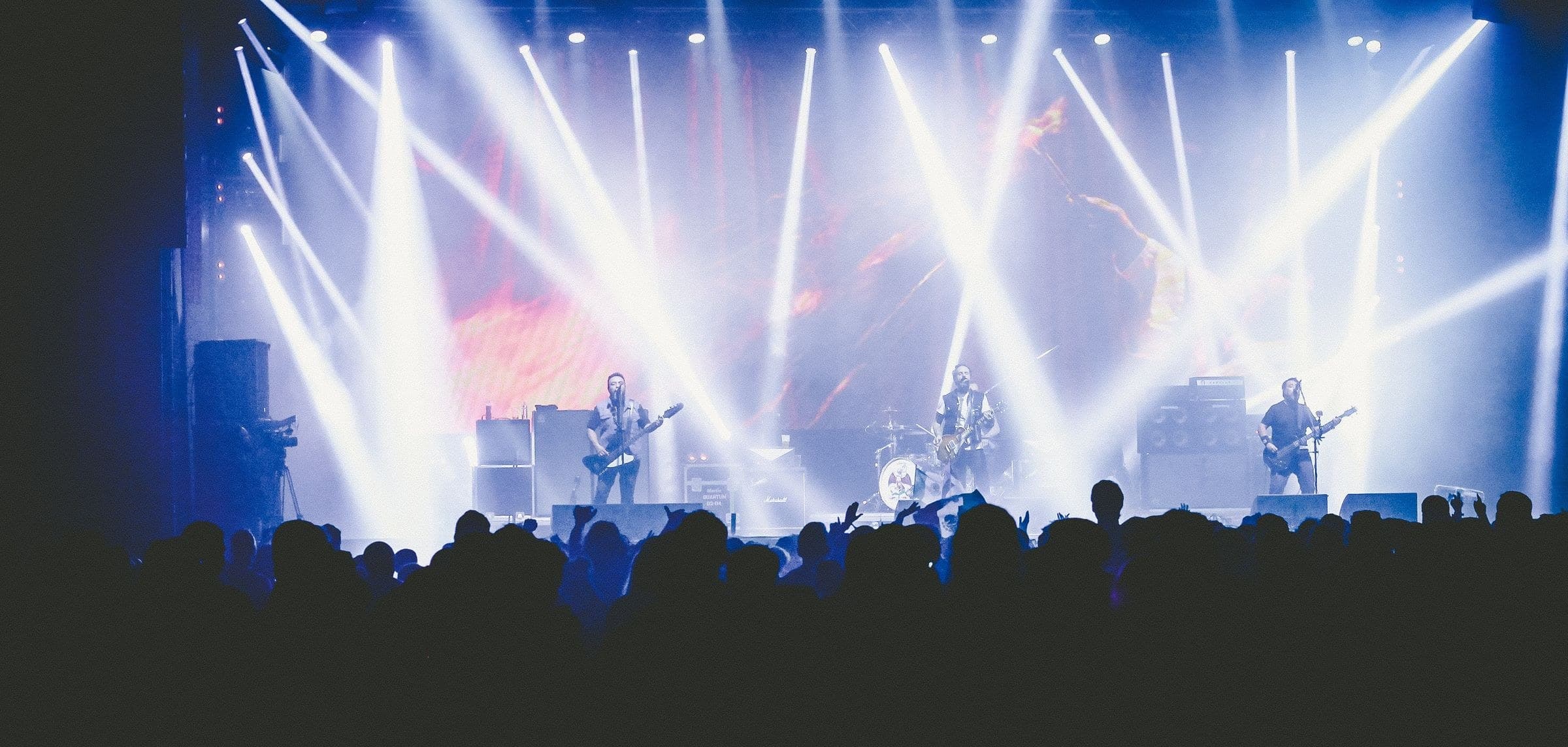 A band performing on a large stage under crossing white light beams with a silhouetted crowd in the foreground