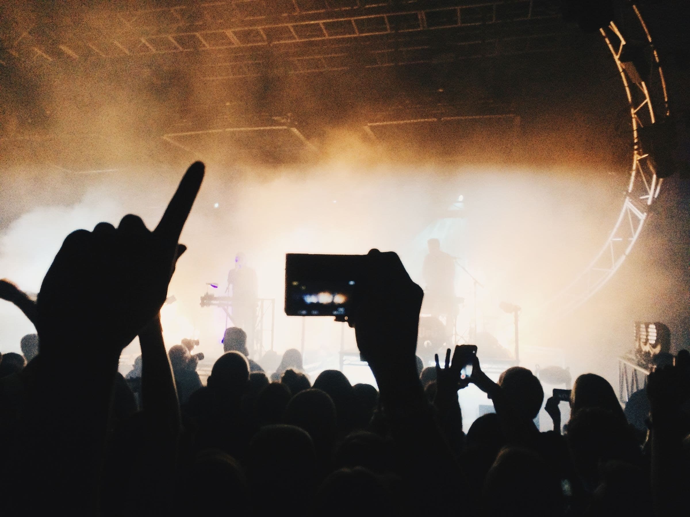 A concert crowd filming the stage on their phones with the band lit by smoky backlight