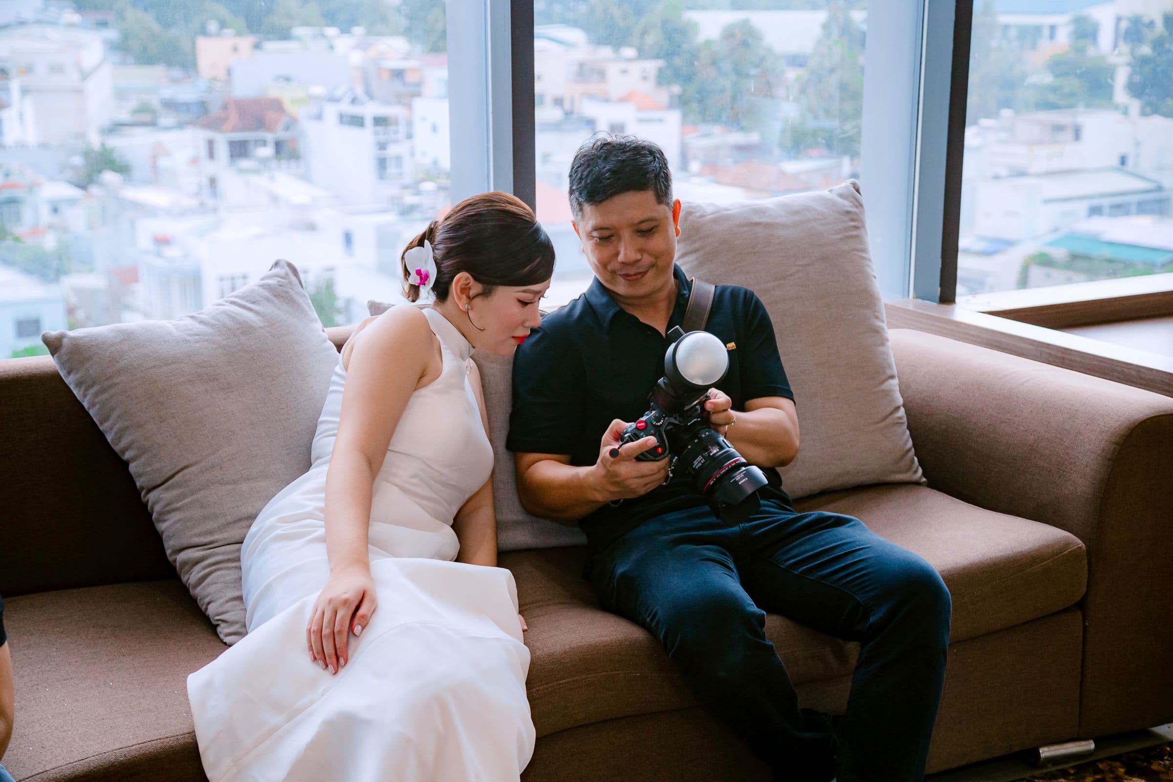 A newly married couple sitting together on a sofa, reviewing their wedding photos on the back of a camera with their photographer