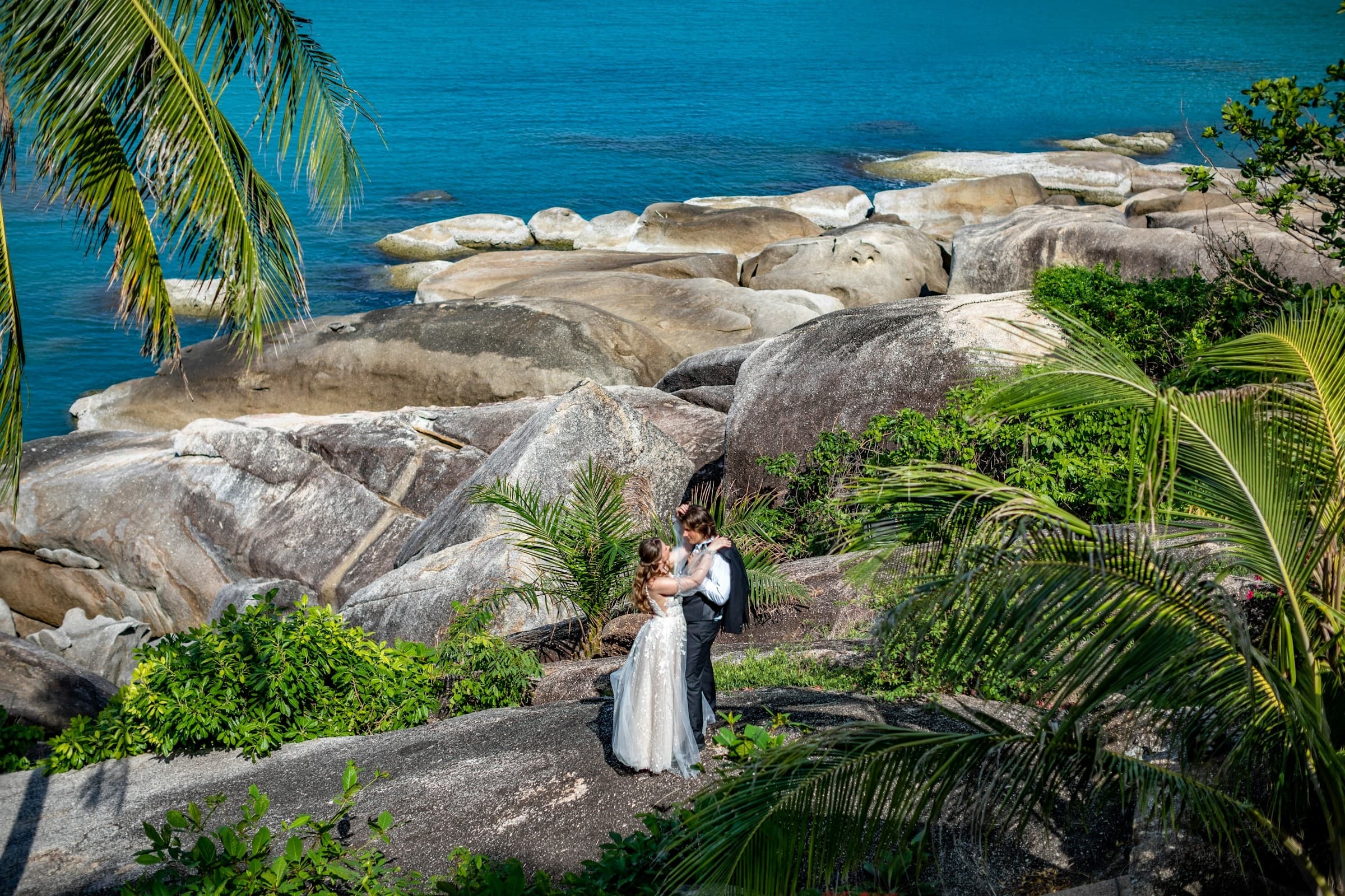 A bride and groom standing together on rocks above a turquoise sea at a tropical destination wedding
