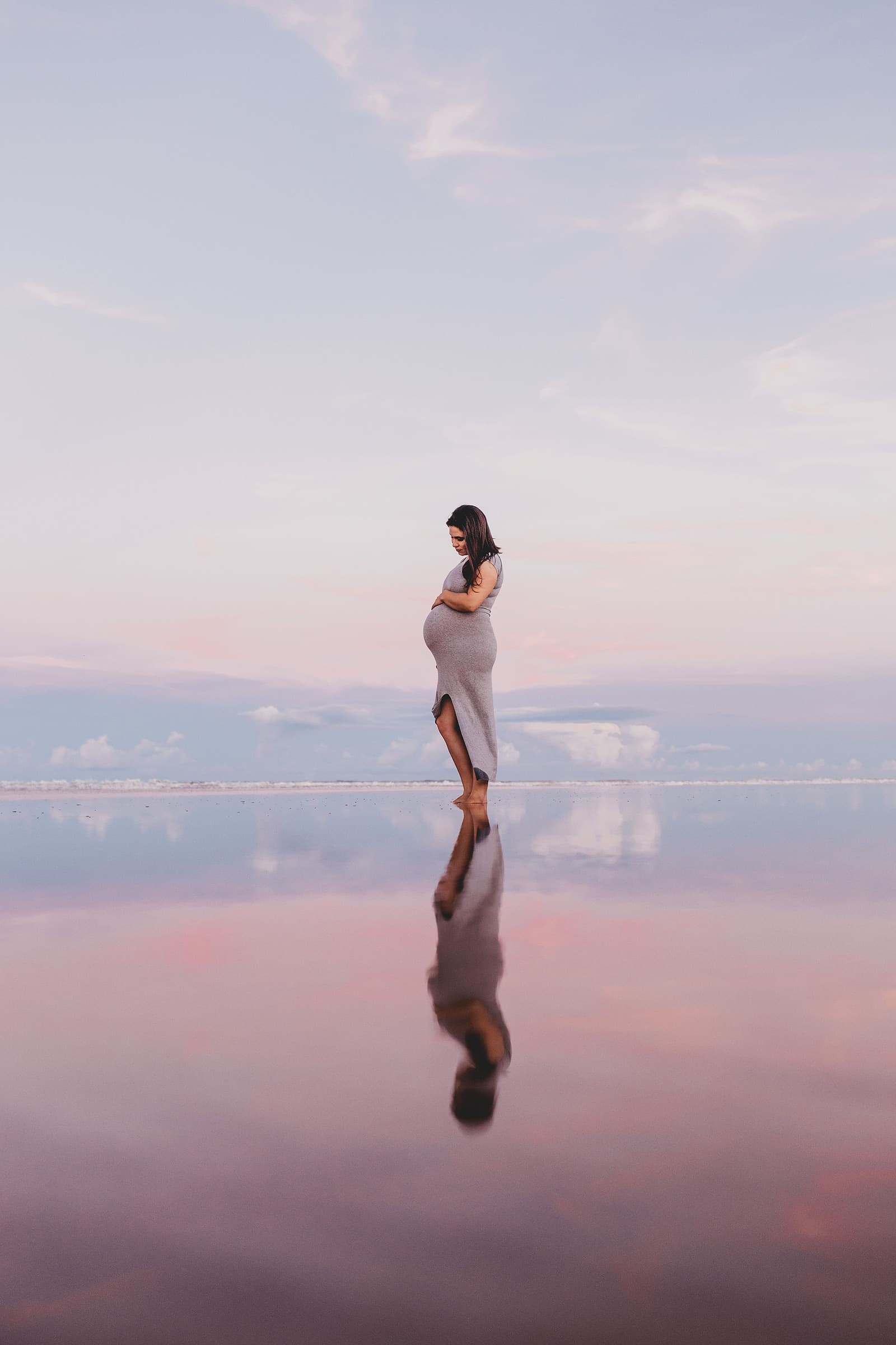A pregnant woman standing in a long dress on a reflective shoreline at sunset during a maternity session