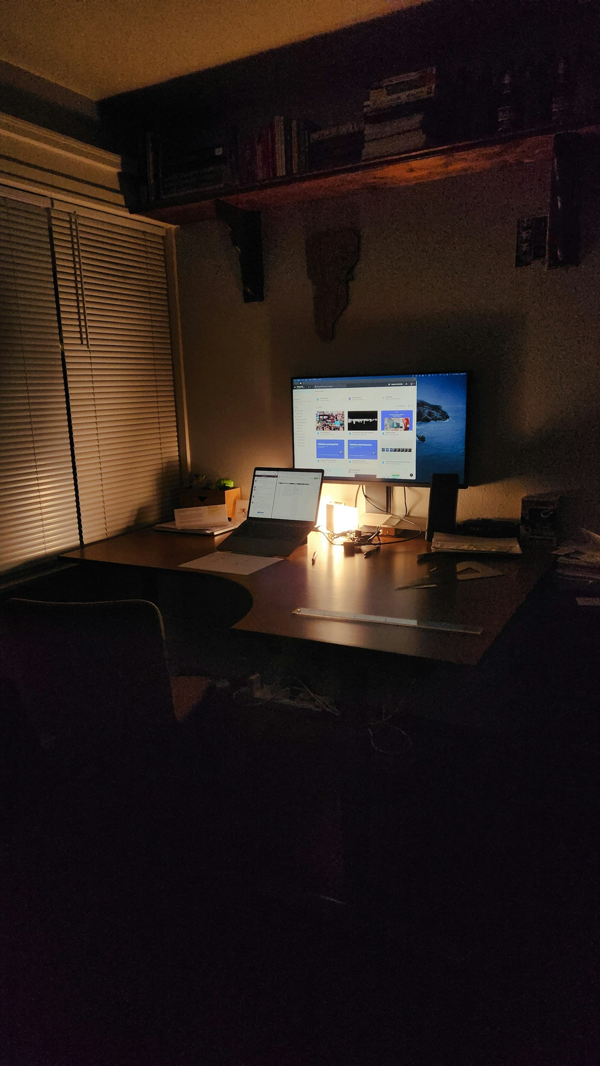 A home desk setup at night with a laptop and monitor lit by a warm desk lamp