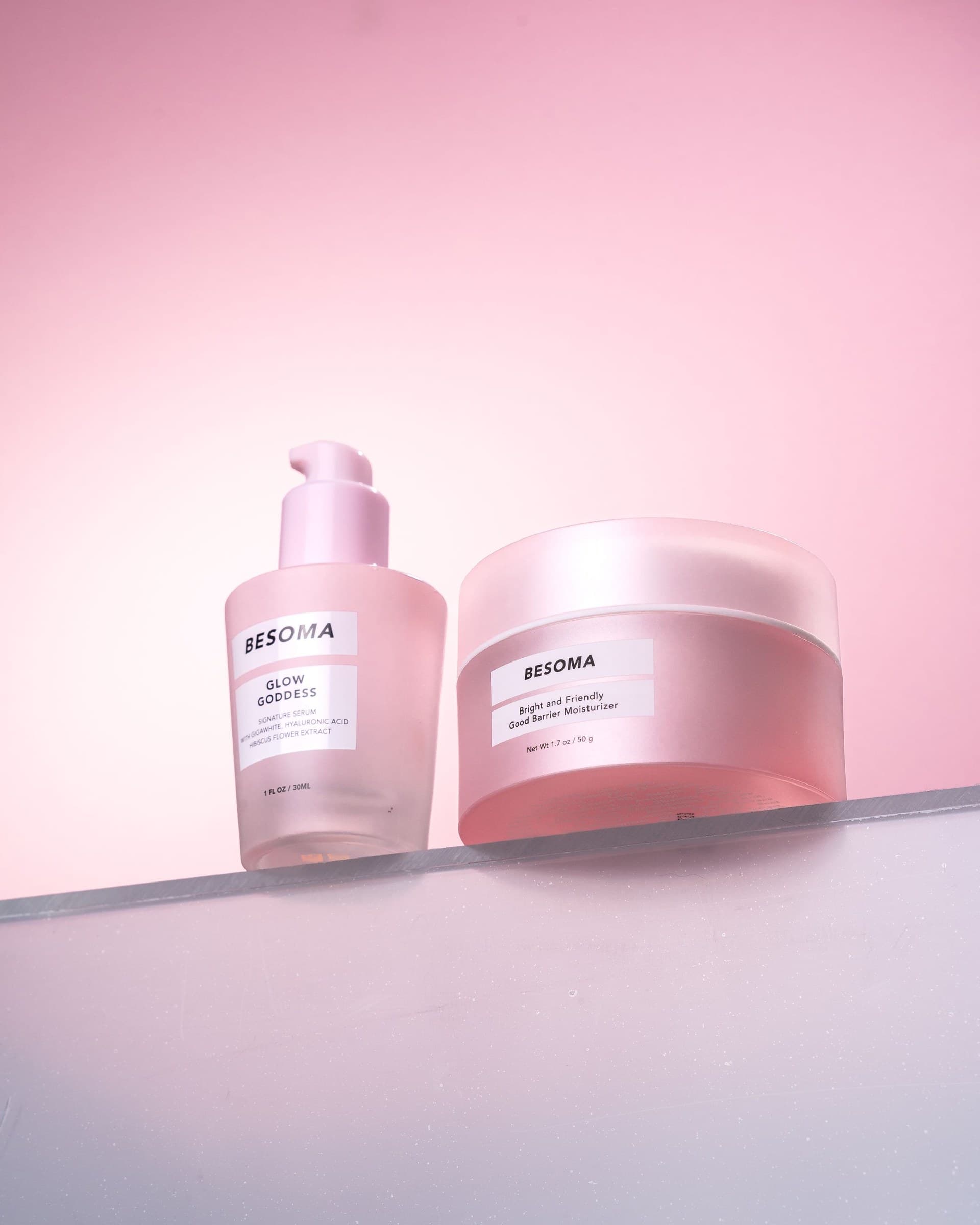 Two frosted pink skincare products styled on a reflective surface against a soft pink gradient background