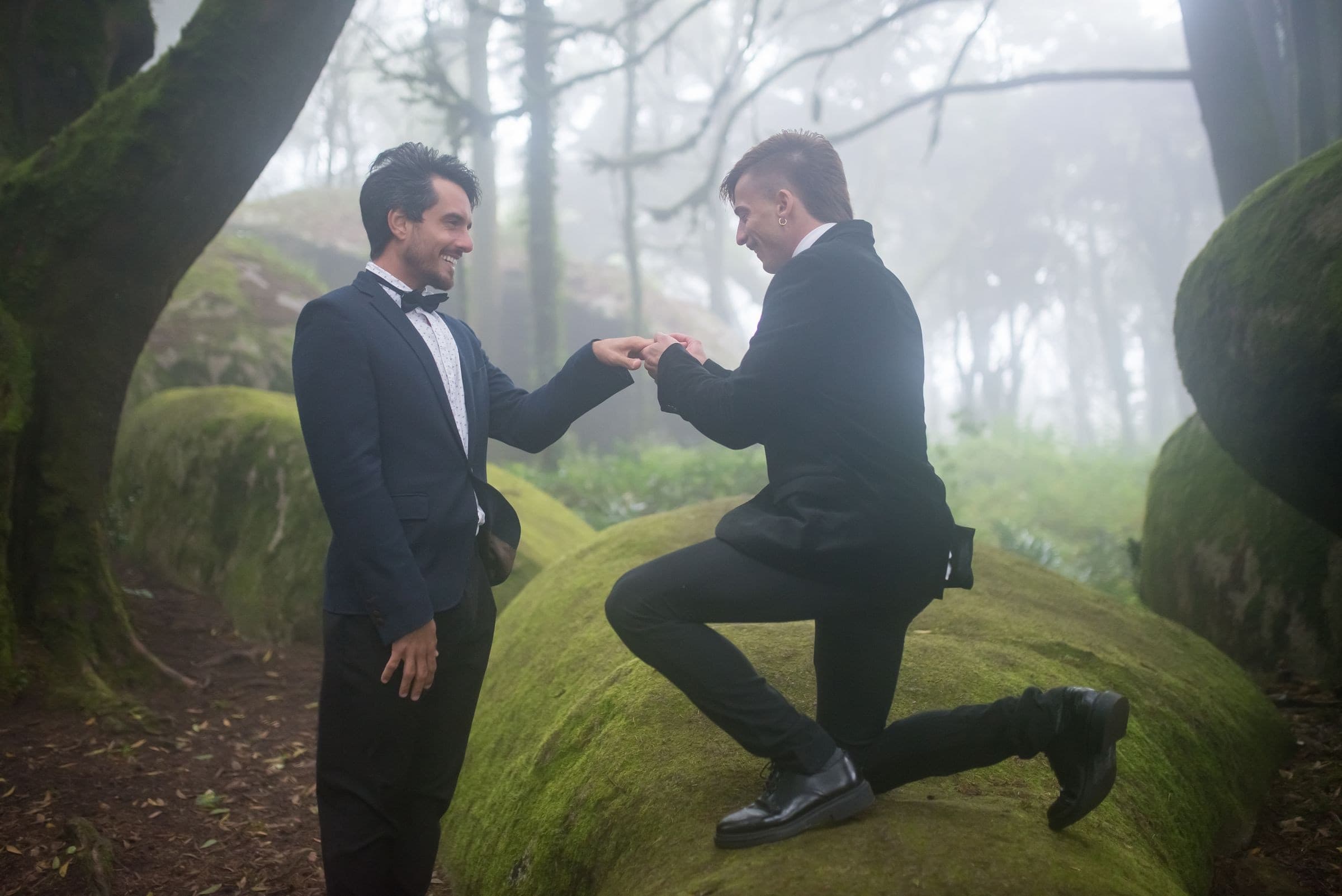 One partner kneeling to put a ring on the other's hand in a misty forest