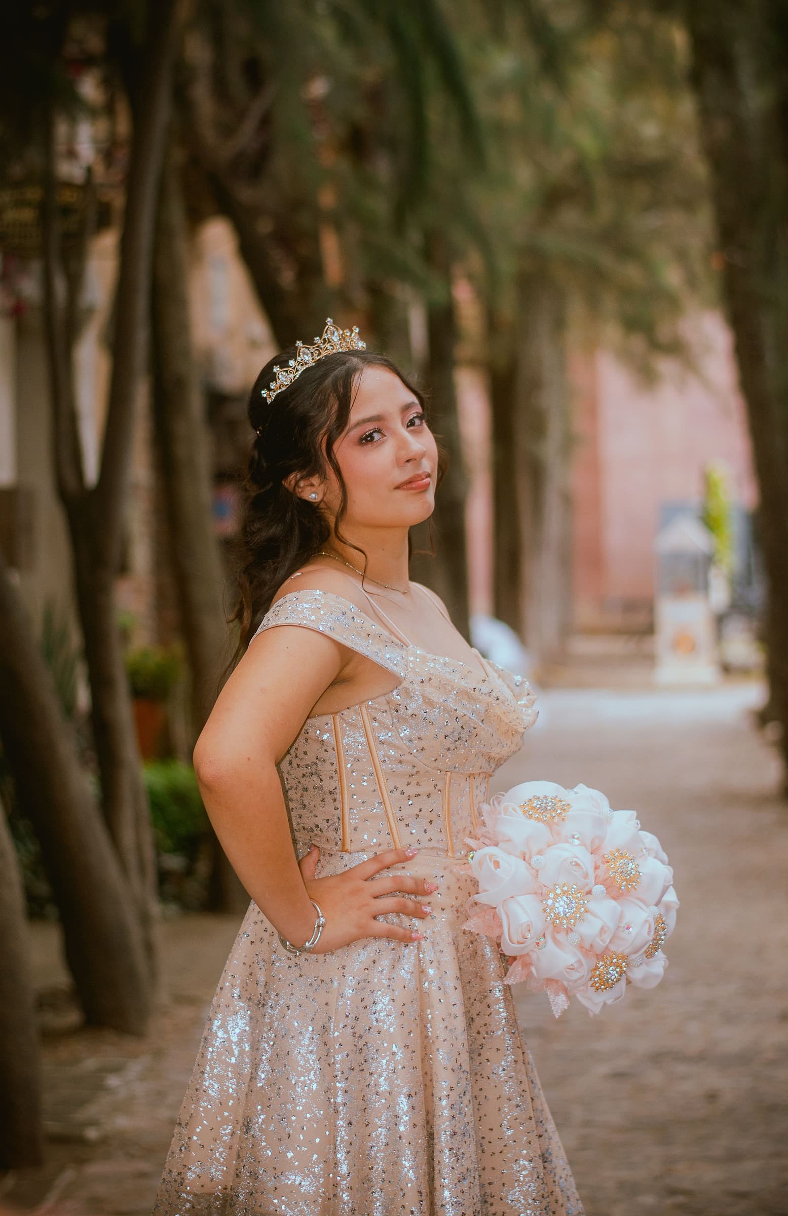 A quinceañera in a champagne sequined ball gown and tiara holding a bouquet on a tree lined path