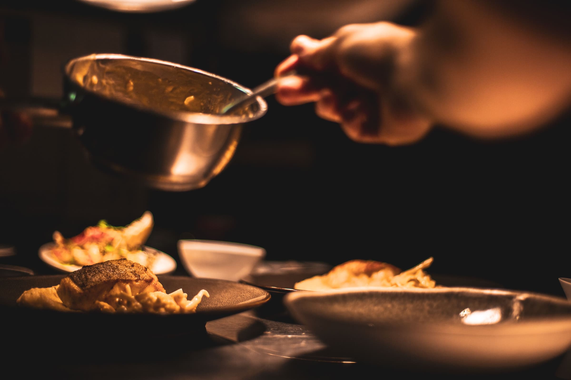 A chef spooning sauce onto a plate in a dark restaurant kitchen