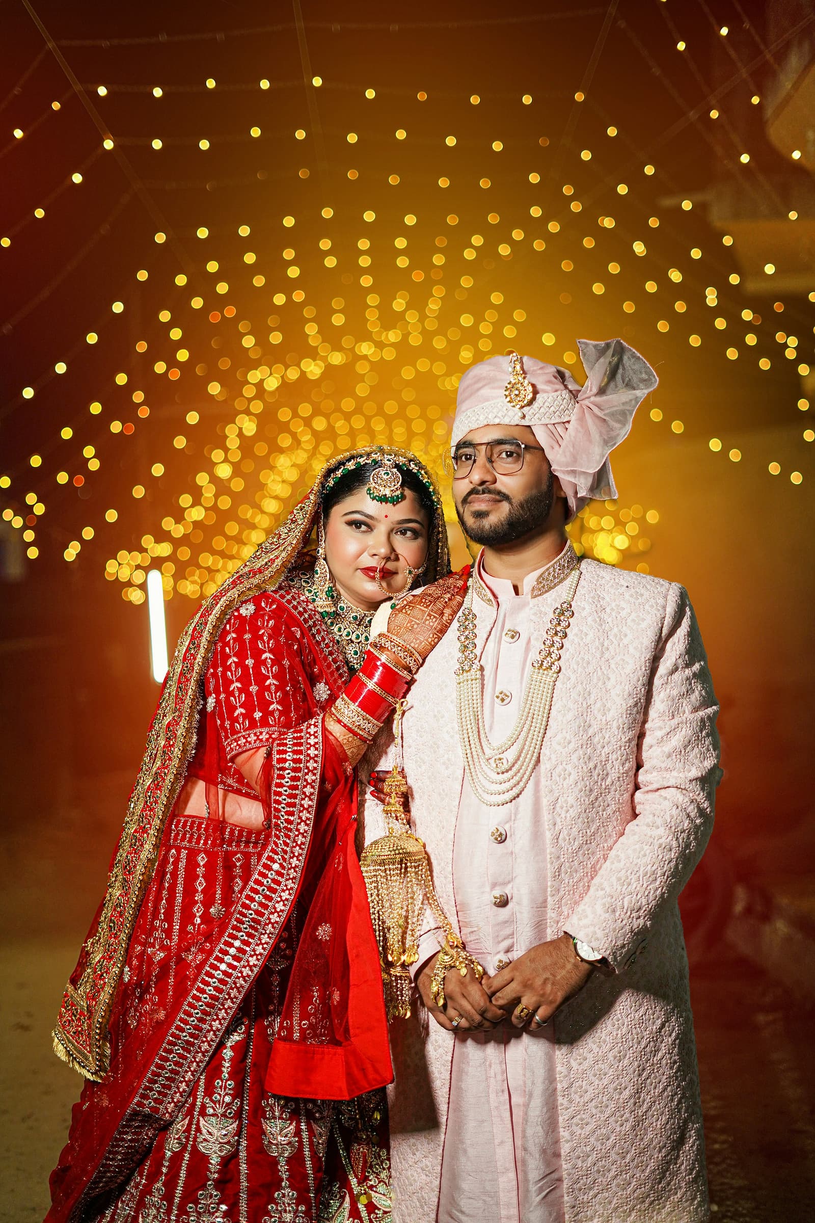 A bride and groom in traditional red and gold wedding dress standing beneath warm string lights