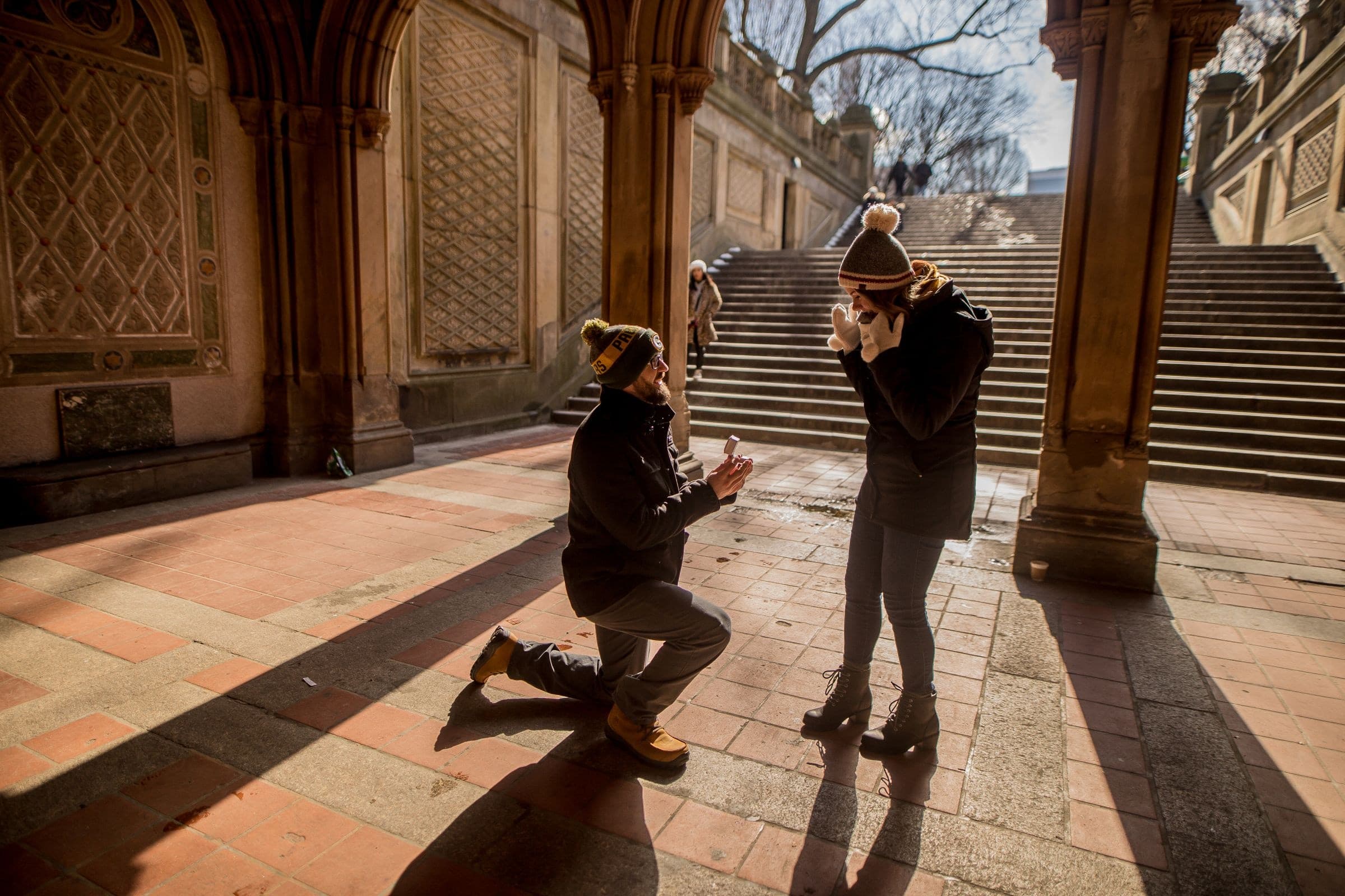 A man kneeling to propose while his partner reacts, photographed from a distance in winter light