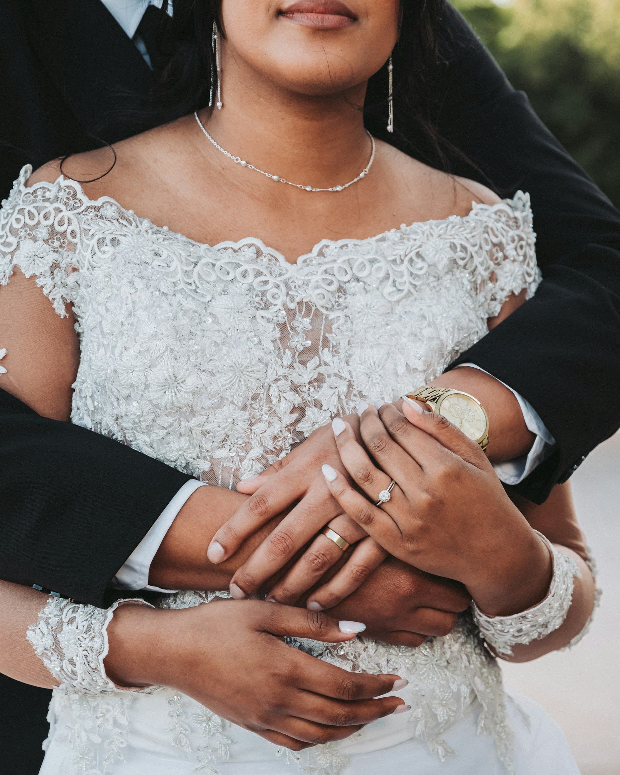 Close-up of a bride and groom's hands, wedding rings visible, as the groom embraces the bride from behind