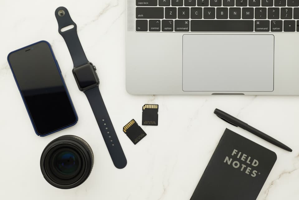 A flatlay of a laptop, phone, camera lens, and dual memory cards laid out on a desk