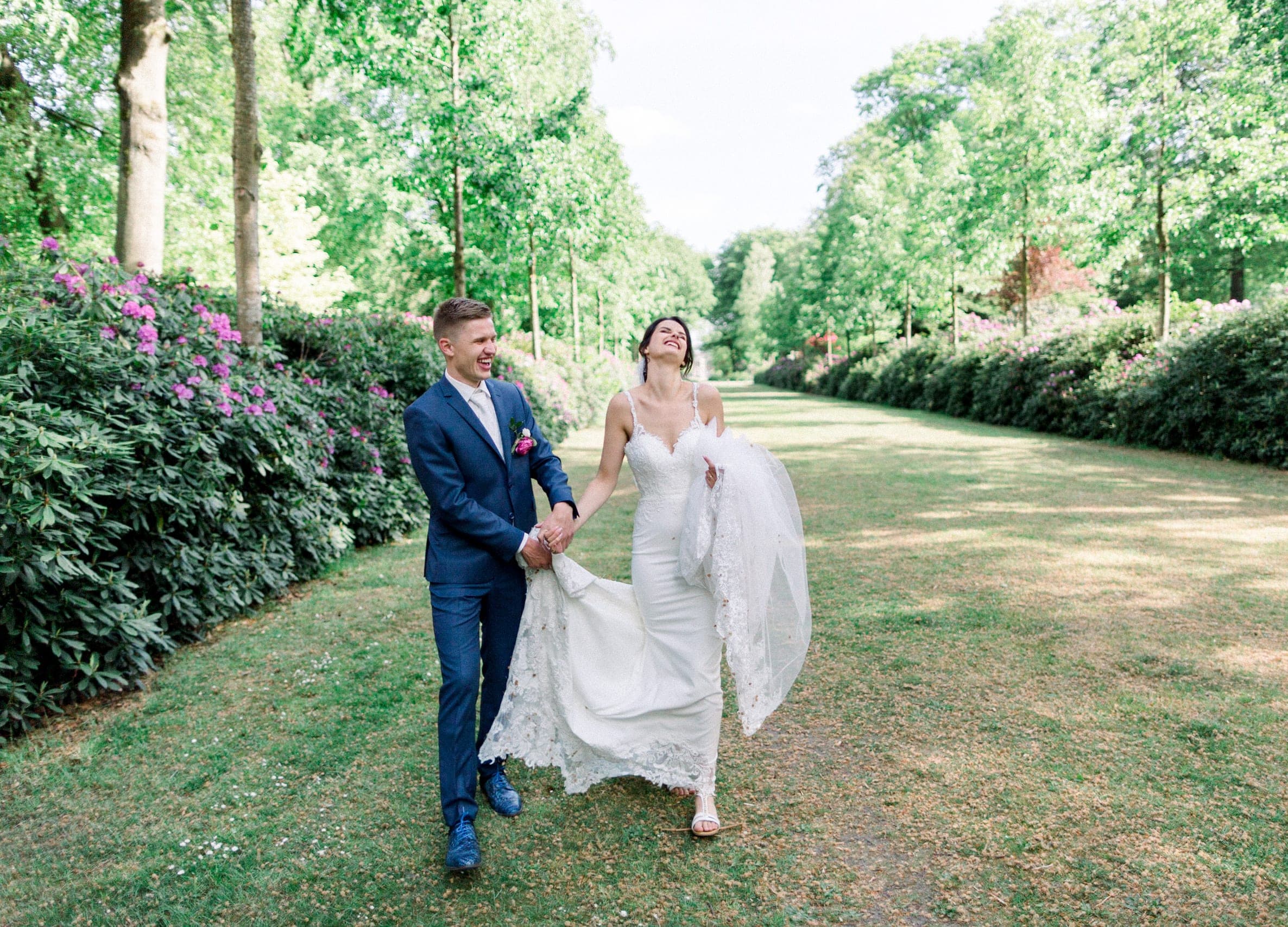 A bride laughing as her groom holds her train while they walk down a garden path lined with flowering hedges